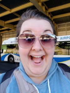 Megan wears a pair of sunglasses and has an excited look on her face. She stands in front of a bus in front of an airport.