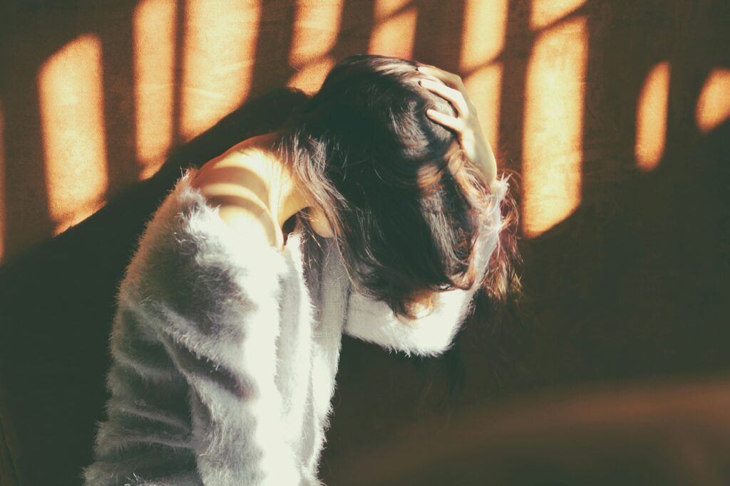 A girl wearing a fuzzy sweater sits in profile has her head bowed and her hand over her head. The fading sun light is coming in through a window.